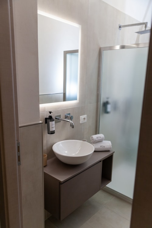 Bagno moderno dell'hotel Marsala con lavabo circolare bianco, specchio illuminato, doccia a pioggia e design minimalista