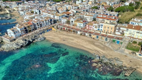 Vista aèria de la costa de Palafrugell amb aigües turqueses del Mediterrani, platja de sorra i edificis històrics de colors vibrants