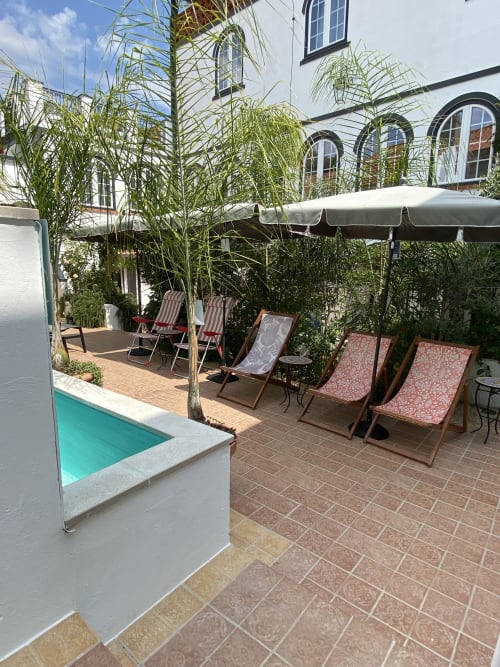Sunny courtyard at Patio Da Villa guesthouse with pool, lounge chairs, palm trees, umbrella, and white historic building in