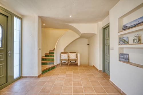 Spacious cottage interior in Palafrugell with terracotta tiles, curved archway, green doors, shelving, and staircase access