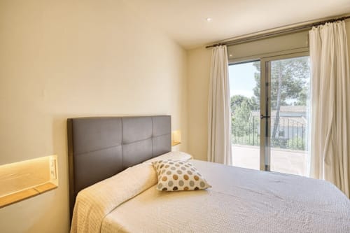 Bright bedroom with gray upholstered headboard, cream bedding, garden view doors, and yellow walls in Palafrugell cottage