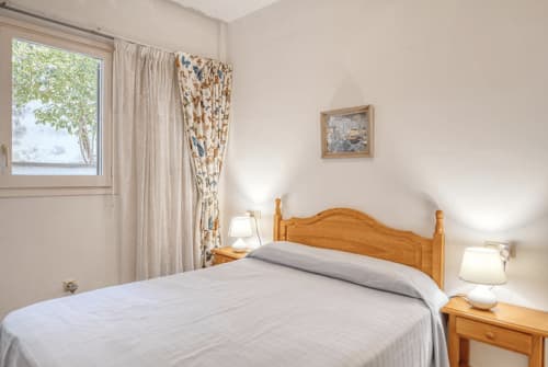 Bright bedroom with wooden bed, white linens, bedside lamps, patterned curtains, and window in Palafrugell apartment