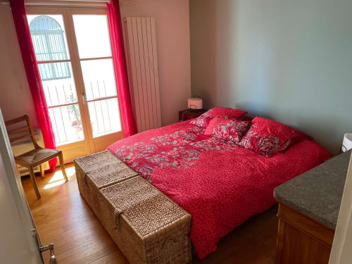 Helles Schlafzimmer mit roter gemusterter Bettwäsche, Holztüren zur Terrasse und neutralen Wänden in La Conciergerie De Franck