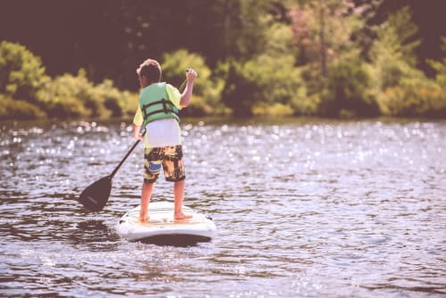 Child paddleboarding on calm water wearing life jacket, forested shoreline behind, Dunoon area activities
