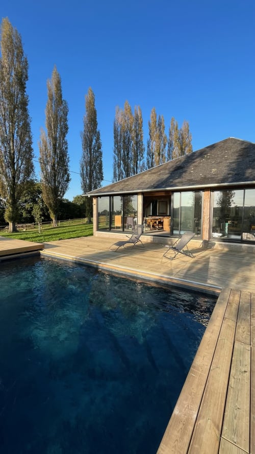 Terrasse piscine moderne du Domaine de Blanvatel avec terrasse en bois, piscine à débordement et peupliers sous ciel dégagé