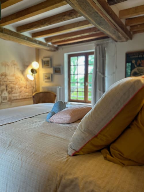 Chambre douillette au Domaine de Blanvatel avec poutres apparentes, lits garnis de coussins et portes-fenêtres sur le jardin