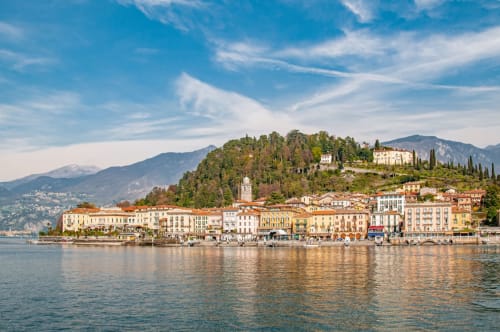 La pittoresca cittadina lacustre di Erba con edifici colorati, montagne e cipressi riflessi nelle acque azzurre
