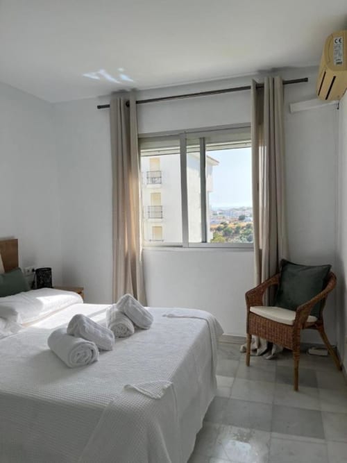 Dormitorio luminoso en Kandy Apartment de Mijas con vistas al mar, ropa de cama blanca, toallas enrolladas y silla de mimbre