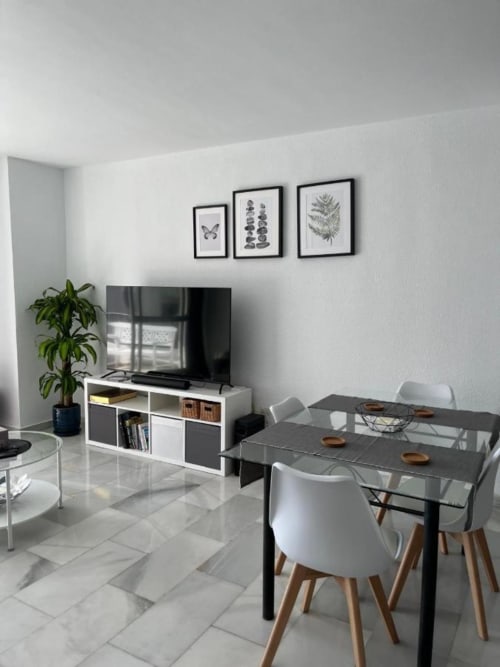 Sala de estar y comedor del apartamento moderno Kandy en Mijas con mesa negra, sillas blancas, mueble TV y arte botánico en la pared