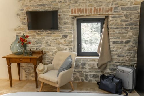 Chambre du gite Amande avec un fauteuil, une table, un mur en pierre, une fenêtre et une télévision. Il y a également un vase de fleurs et un chapeau sur le fauteuil.
