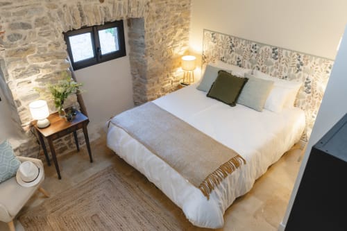 Une chambre rustique avec un lit double, des draps blancs et une couverture en laine. Le mur est en pierre et une fenêtre donne sur l'extérieur. Il y a également des lampes de chevet, une table de nuit et un fauteuil.