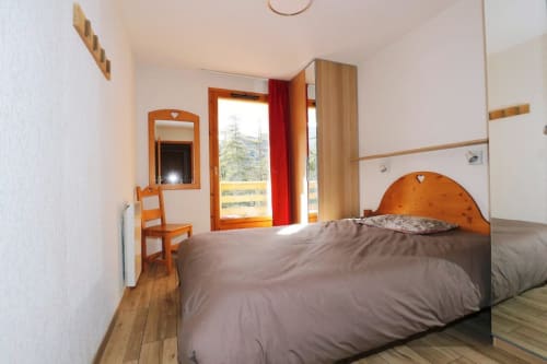 "Chambre cosy de l'appartement 301 à Vars-les-Claux, avec un sommier en bois élégant, offrant un cadre chaleureux et confortable pour une bonne nuit de repos après une journée de ski."