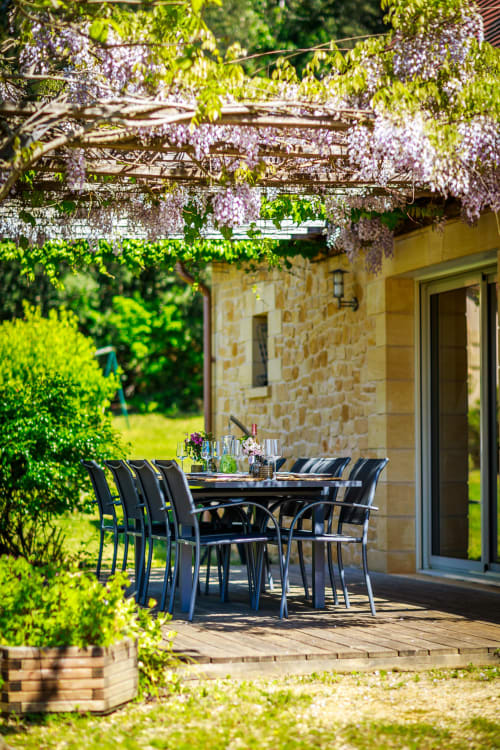 Openluchteetplaats op het terras van Domaine De Campagnac met zwarte stoelen, blauweregen-pergola en stenen cottage in Carsac-Aillac
