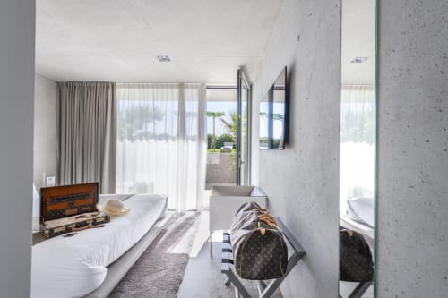Modernes Schlafzimmer im Le Pavillon M mit weißem Bett, Betonwänden, großen Fenstern mit Blick auf Grignan und Designer-Gepäck.