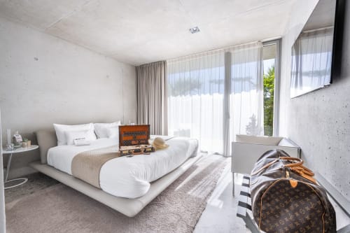 Modernes Schlafzimmer im Le Pavillon M in Grignan mit weißem Bett, Betonwänden, großen Fenstern und Designer-Gepäck.