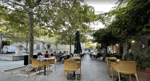 Yellow chairs and tables at AJOUCADOU's outdoor terrace in Lourmarin, surrounded by trees and historic buildings