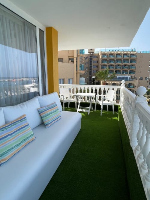 Modern balcony with white lounge, striped pillows, artificial grass, white railing, ocean and neighboring buildings at Sunset