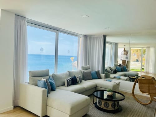 Sunset ocean view apartment living room with cream sectional, blue cushions, and floor-to-ceiling windows overlooking the sea