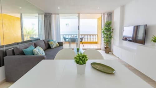 Modern 1-bedroom apartment in Arguineguín with sea view balcony, gray sofa, white dining table, and flat-screen TV