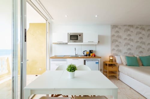 Modern studio apartment kitchen with sea view balcony, white cabinetry, wooden countertop, and bed with ocean backdrop in