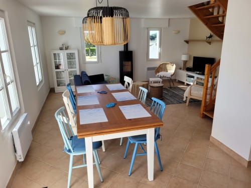 Spacieuse salle à manger et salon ouvert dans le cottage Belle Île avec table en bois, chaises bleues, cheminée et vue sur le jardin