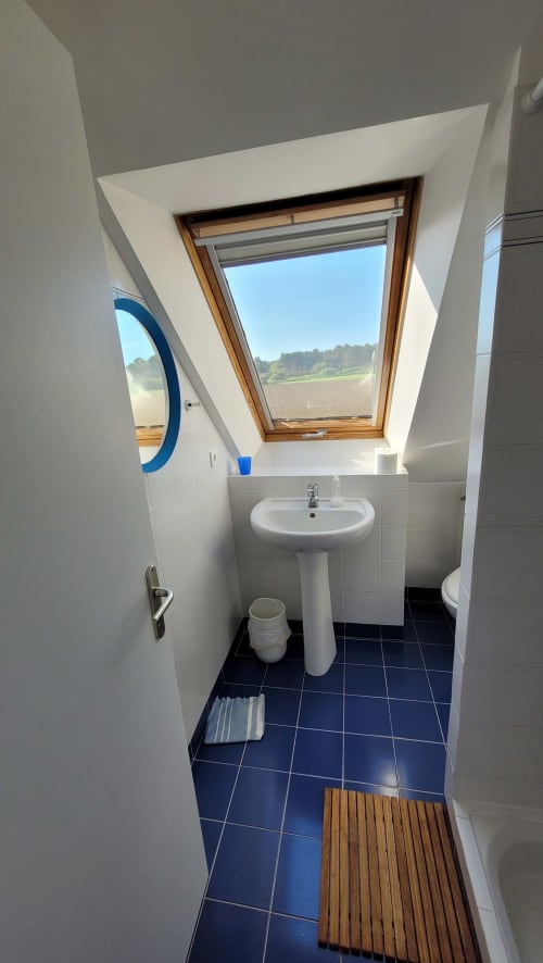 Salle de bain moderne avec puits de lumière donnant sur la campagne de Belle Île, lavabo sur piédestal et carrelage bleu foncé à Locmaria