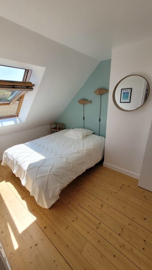 Chambre mansardée lumineuse avec lit blanc, mur vert sage, puits de lumière, miroir rond et parquet à Belle Île en Mer