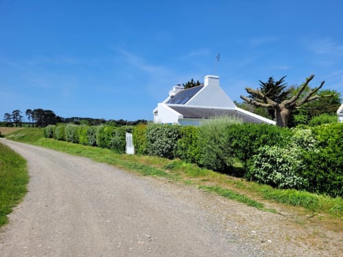 Maison blanche avec panneaux solaires à Belle-Île-en-Mer, entourée de haies verdoyantes et allée de gravier sous un ciel bleu