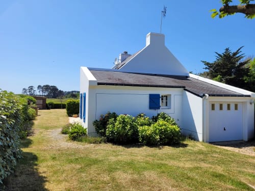 Maison blanche aux volets bleus et toit pointu caractéristique sur Belle Île en Mer, entourée de pelouse verte et d'arbustes de jardin