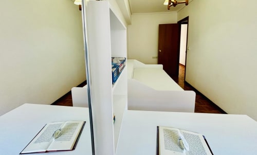 Dormitorio compartido con dos camas separadas por divisores blancos en el apartamento Breixo, Baiona. Libros abiertos en escritorios con vistas a la ciudad
