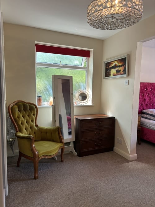 Elegant bedroom seating area with olive velvet chair, dark wood dresser, and garden view at England Furnished Homes luxury