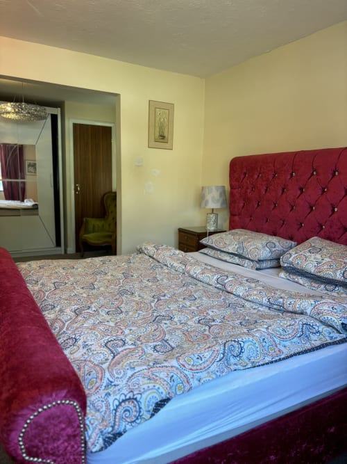 Spacious bedroom with burgundy tufted headboard, patterned bedding, lamp, and yellow walls at England Furnished Homes villa