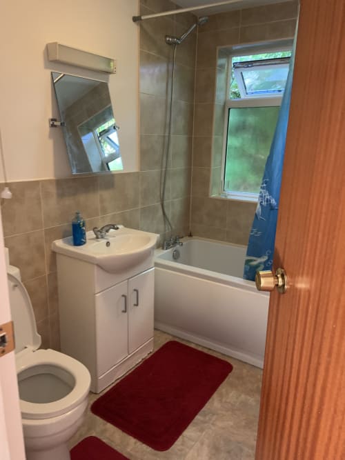 Modern bathroom with white sink vanity, toilet, bathtub with shower screen, tiled walls, mirror, and red bath mat in England