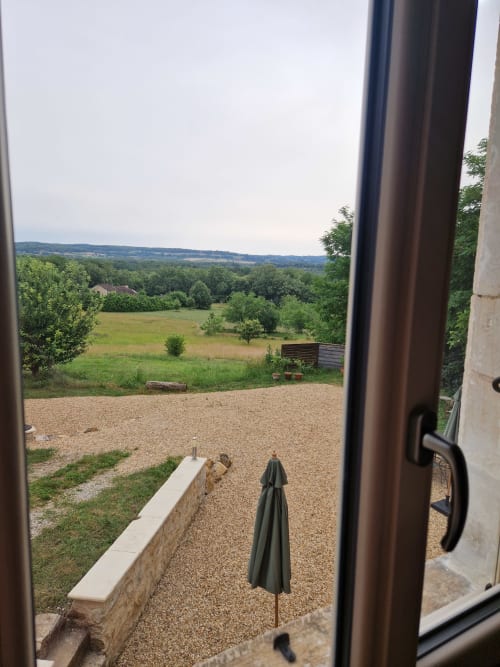 Vue du jardin Maisonaurinko à Thénon avec parapluie vert sur terrasse en gravier dominant la campagne vallonnée