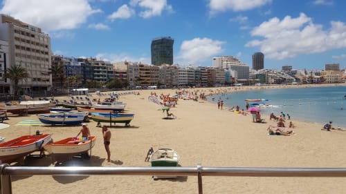 Arguineguín beach with fishing boats, sandy shore, swimmers, and coastal buildings under blue sky