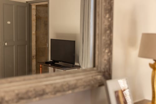 Madeleine Room at Le Prieuré Notre-Dame showing flat-screen TV on stand, mirrored wardrobe, and curtained window in Montfrin