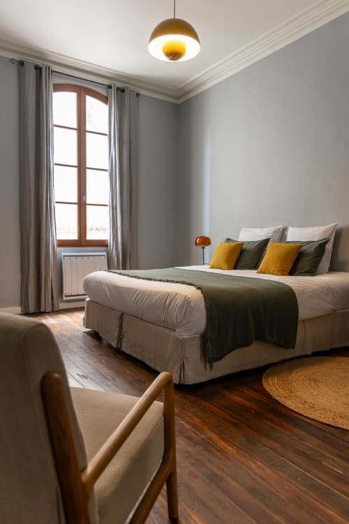 Chambre moderne du Prieuré Notre-Dame avec plafond à poutres apparentes, suspension dorée et vue sur les monuments de Montfrin