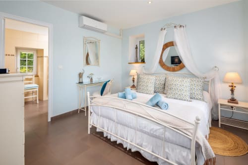 Lichte blauwe slaapkamer met wit ijzeren bed, bloemenprint kussens en witte baldakijn gordijnen in cottage Domaine De Campagnac