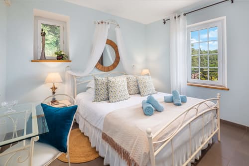 Heldere slaapkamer in gîte Domaine De Campagnac met wit ijzeren bed, blauwe accenten, houten spiegeldosel en tuinzicht