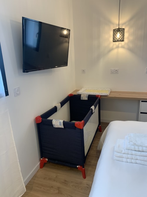 Camera da letto moderna con TV a parete, lettino portatile con accenti rossi, biancheria bianca, luce a sospensione e pavimento in legno