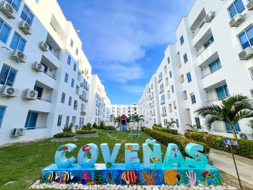 Complejo residencial Coveñas con colorido letrero COVEÑAS en jardín, edificios modernos blancos con acentos azules y palmeras