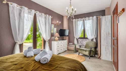 Elegante slaapkamer in Domaine De Campagnac met gouden beddengoed, witte gordijnen, kroonluchter, TV en tuinuitzicht