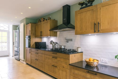 Cuisine contemporaine avec meubles en bois, plan de travail noir, dosseret en carreaux blancs et cuisinière à gaz au Prieuré Du Preux