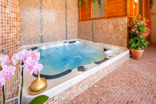Luxurious hot tub with blue water surrounded by terracotta tile, pink orchids, and wooden door at Domaine De Campagnac