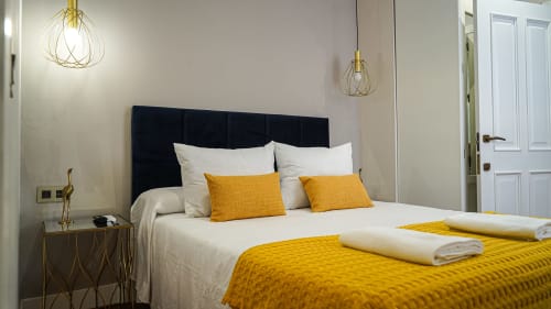 Moderno dormitorio en apartamento Gestyp-AT en Mérida con cabecero negro, lámparas doradas, detalles amarillos y vista a la ciudad