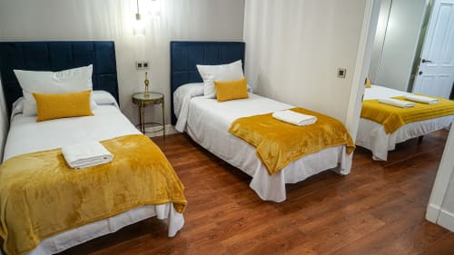 Dormitorio moderno en Gestyp-AT Mérida con tres camas, cabeceros azul marino, detalles amarillos y piso de madera.