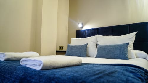 Dormitorio moderno en Mérida con cabecera azul marino, ropa blanca, almohadas azules, toallas enrolladas e iluminación de ambiente