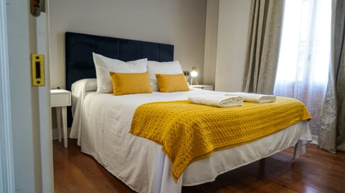 Dormitorio moderno en apartamento de Mérida con cabecera negra, ropa de cama blanca, detalles dorados y piso de madera.