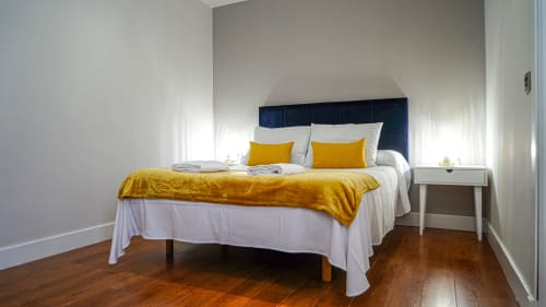 Dormitorio moderno en apartamento Gestyp-AT en Mérida con cabecera azul marino, detalles amarillos, ropa de cama blanca y piso de madera