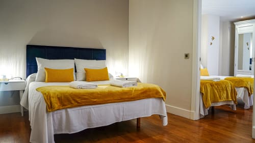 Dormitorio luminoso en Mérida con cabecero azul marino, ropa de cama blanca y acentos de terciopelo dorado con vista a la ciudad